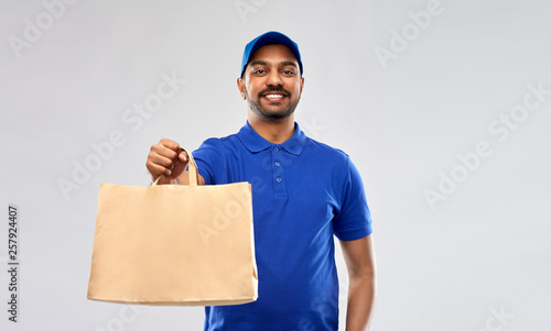 service and people concept - happy indian delivery man food in paper bag in blue uniform over grey background