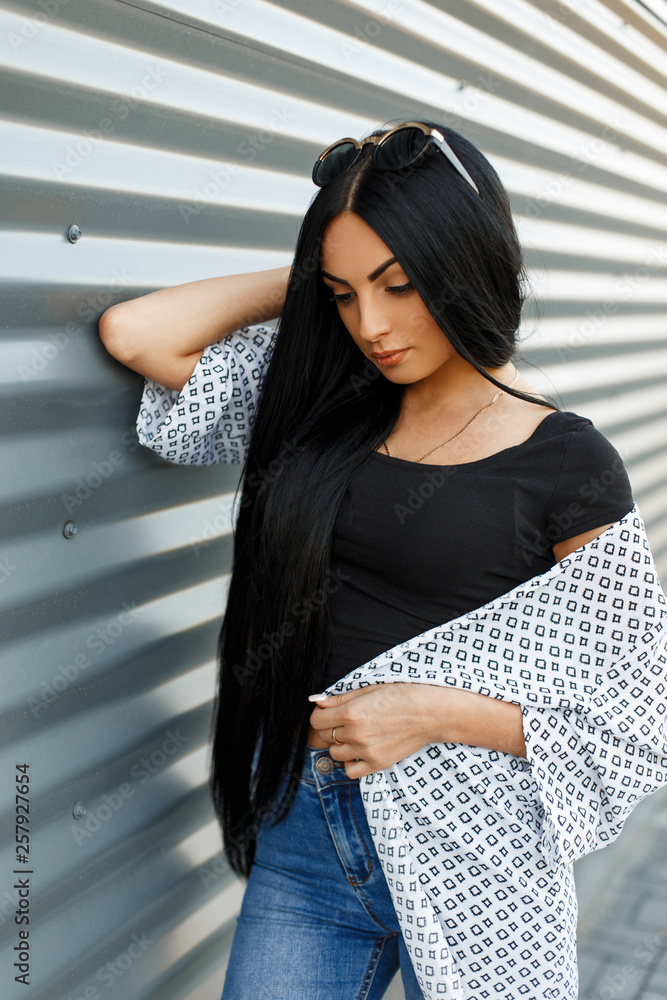 Fashionable european young hipster woman with long luxurious hair in a black  top in a jacket in vintage jeans posing standing near a metallic striped  wall outdoors. Sexy girl model. Stock Photo |