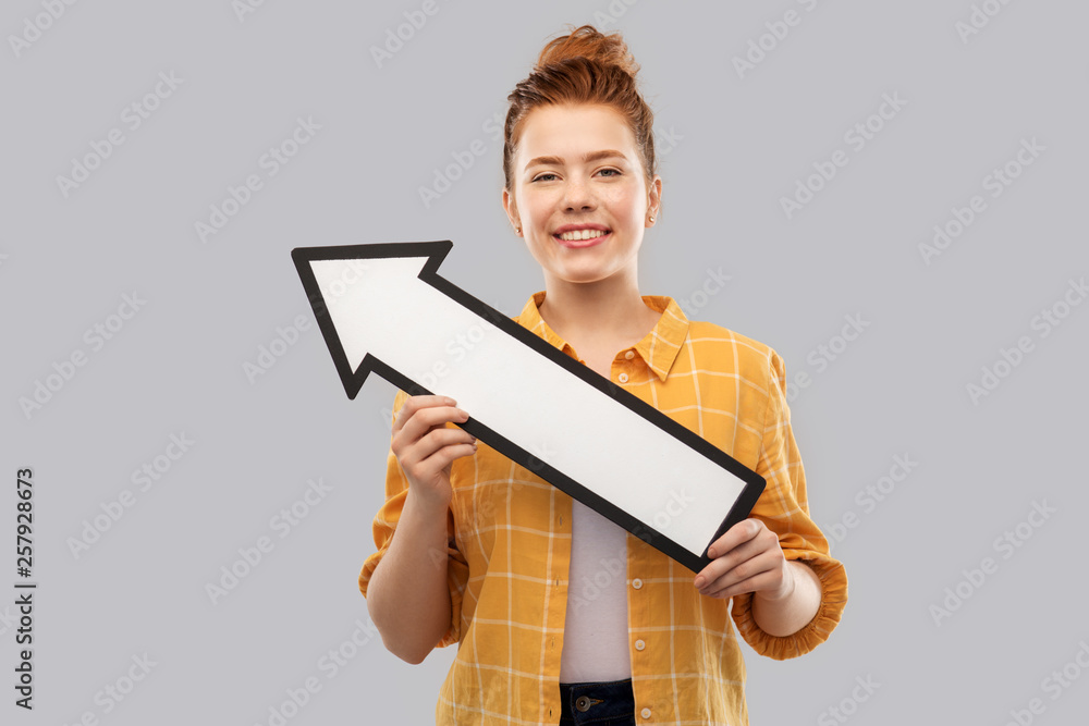 people concept - smiling red haired teenage girl in checkered shirt holding big white thick arrow showing to north west over grey background people concept - smiling red haired teenage girl in checkered shirt holding big white thick arrow showing to north west over grey background