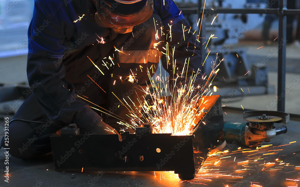 The worker sits on the concrete floor in the workshop and welded metal part. Fly bright sparks.