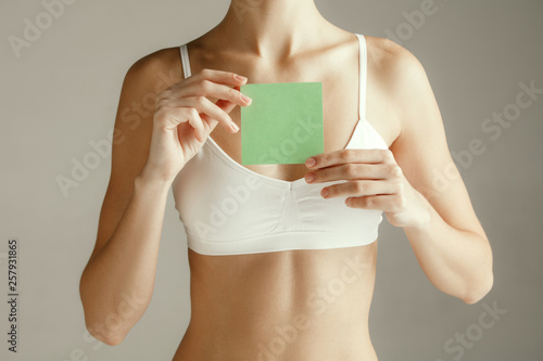 Woman health. Female model holding empty card near breast. Young adult girl with paper for sign or symbol isolated on gray studio background. Cut out part of body. Medical problem and solution.