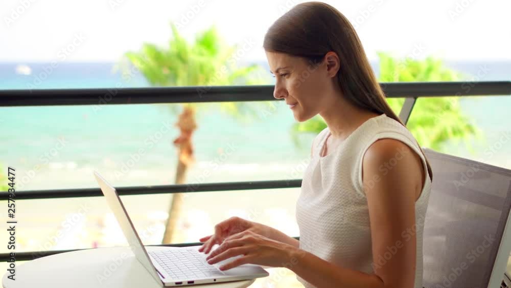 Businesswoman sitting on balcony working on laptop at tropical beach resort. Young professional female freelancer typing on computer. Beautiful turquoise sea and palm trees on background Businesswoman sitting on balcony working on laptop at tropical beach resort. Young professional female freelancer typing on computer. Beautiful turquoise sea and palm trees on background