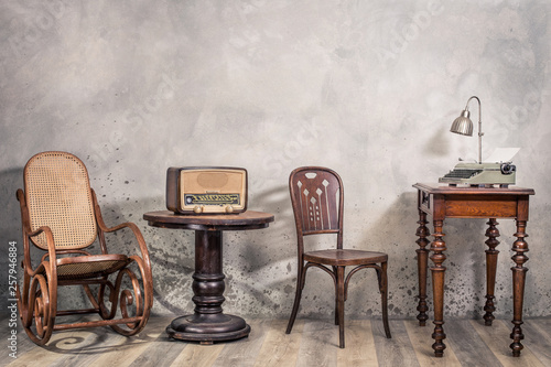 Vintage loft room with antique rocking chair, broadcast radio, old typewriter and lamp on oak wooden desk front concrete wall background with shadows. Retro style filtered photo