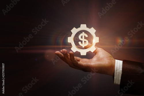 A cogwheel with dollar currency unit inside hanging above the male hand.