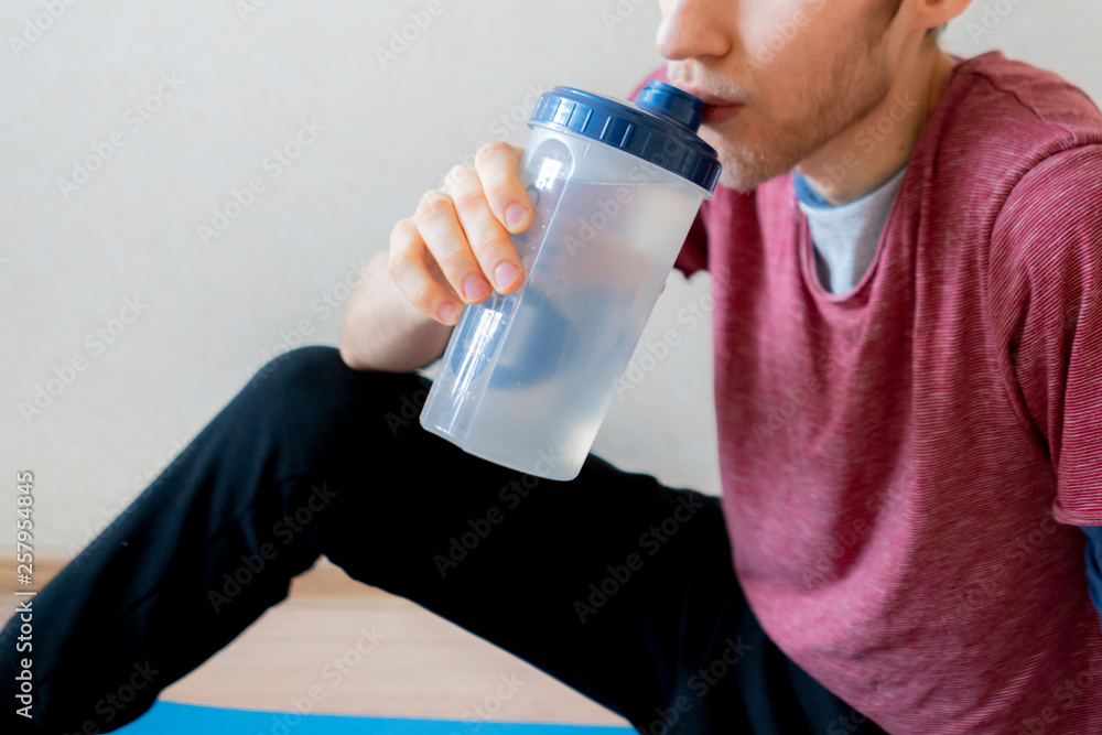 skinny young guy drinking water from shaker while sitting on the floor f skinny young guy drinking water from shaker while sitting on the floor f