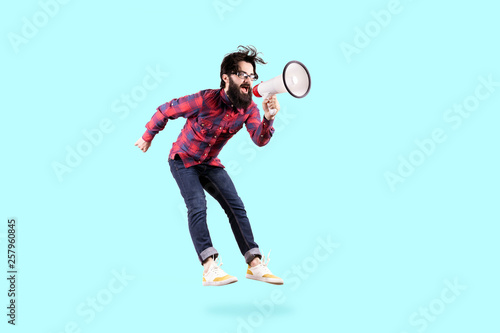 Bearded hipster man in glasses, jumping and shouting in megaphone, photo on blue background © alesmunt