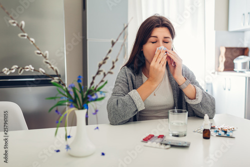 Spring allergy. Young woman sneezing because of flowers surrounded with pills on kitchen. Seasonal allergy concept.