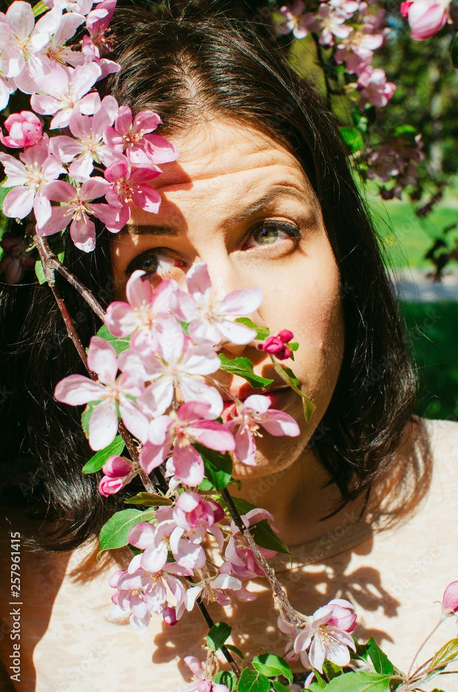 Fototapeta premium brunette girl stands near a blossoming branch of a Apple tree. pink flowers on the face. it's hard to look at the sun, hurts my eyes