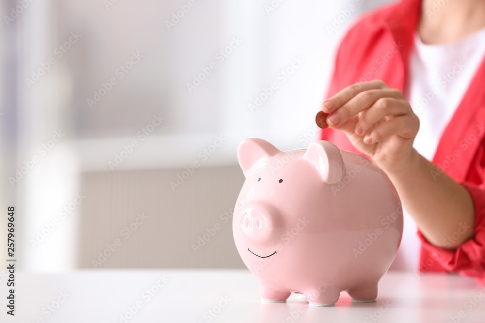 Woman putting coin into piggy bank at table indoors, closeup. Space for text Woman putting coin into piggy bank at table indoors, closeup. Space for text