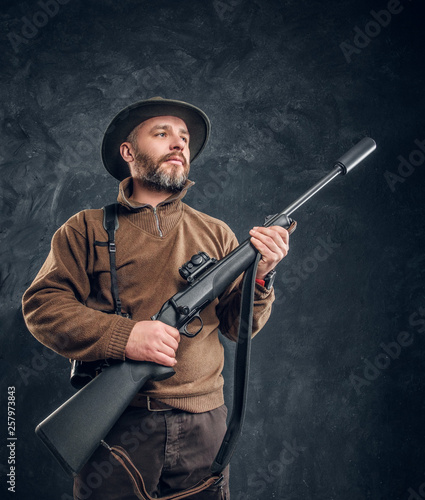 Portrait of a bearded hunter holding a rifle and looking sideways. Studio photo against a dark wall background