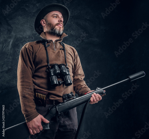 Portrait of a mature hunter with rifle and binoculars. Studio photo against dark wall background