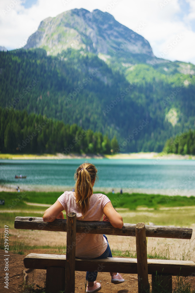 Tourist girl enjoys the magical view sitting on a wooden bench on the shore of a turquoise lake in the mountains. 