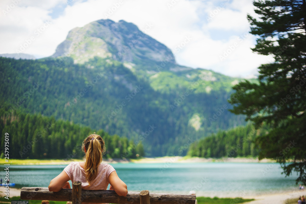 Tourist girl enjoys the magical view sitting on a wooden bench on the shore of a turquoise lake in the mountains. 
