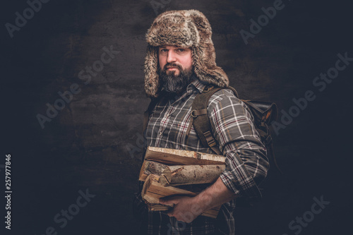 Portrait of a bearded woodcutter with a backpack dressed in a plaid shirt and trapper hat holding firewood. Studio photo against a dark textured wall photo