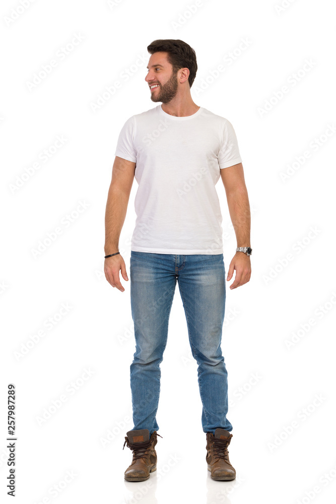 Smiling Man In Jeans And White T-shirt Is Standing And Looking Away. Front View. Smiling Man In Jeans And White T-shirt Is Standing And Looking Away. Front View.