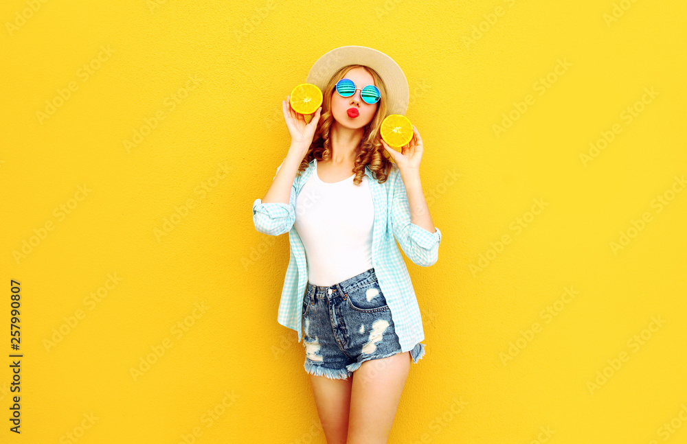 Summer portrait happy woman holding in her hands slices of orange sending sweet air kiss in straw hat, shorts on colorful yellow background Summer portrait happy woman holding in her hands slices of orange sending sweet air kiss in straw hat, shorts on colorful yellow background