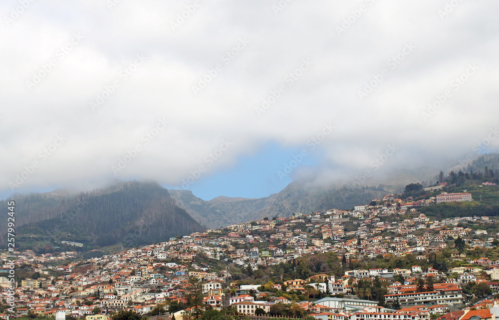 a scenic panoramic aerial cityscape of the city of funchal in Madeira with buildings of the city in front of mountains with the tops covered by white clouds
