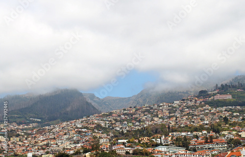 a scenic panoramic aerial cityscape of the city of funchal in Madeira with buildings of the city in front of mountains with the tops covered by white clouds