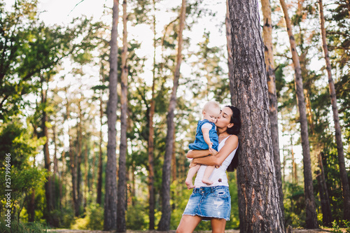 young beautiful fashionable mother with long brunette hair holding a daughter's blonde with blue eyes one year of birth in a coniferous forest in a summer park. Wearing jeans clothes