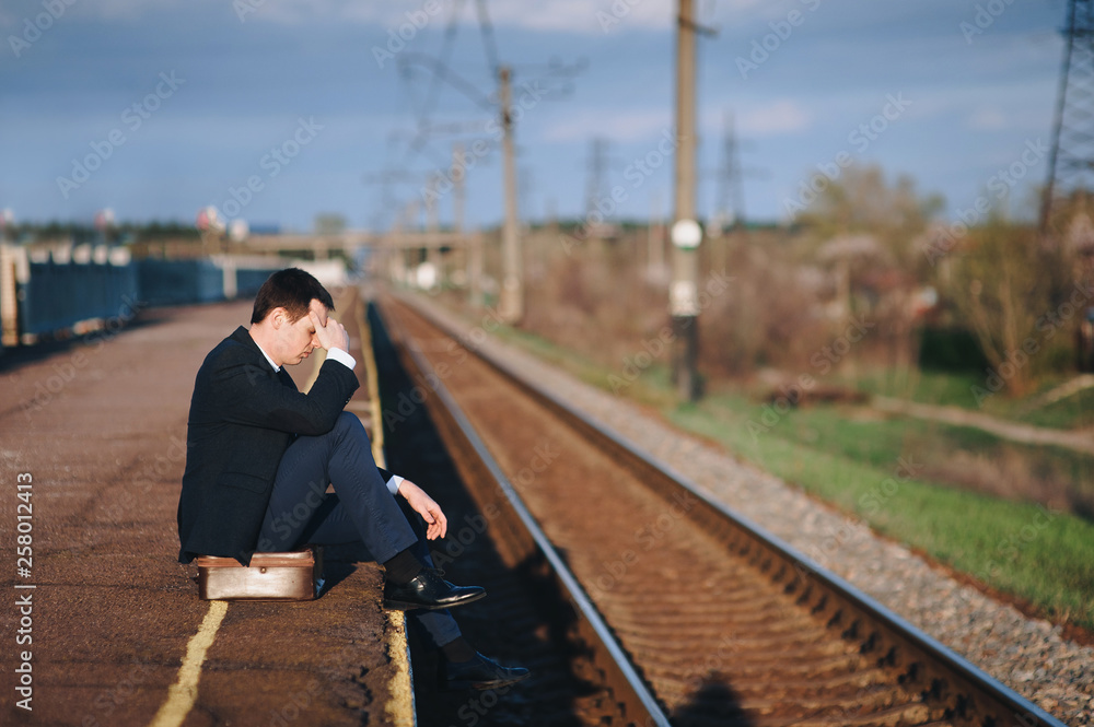 A humiliated and offended businessman is sitting and waiting for the train on the platform. Dismissal, depression and headache. Stylish man in a black suit on the background of clouds and sky.