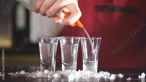 Expert barman in elegant uniform working at night party at popular nightclub, pouring vodka in frosted short glasses photo