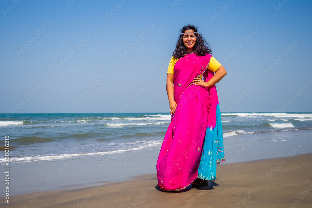 portrait of a beautiful smiling snow-white smile indian woman black curly hair and dark skin in a pink sari holding bottle of sunscreen spray on beach.girl enjoying spf body paradise vacation portrait of a beautiful smiling snow-white smile indian woman black curly hair and dark skin in a pink sari holding bottle of sunscreen spray on beach.girl enjoying spf body paradise vacation