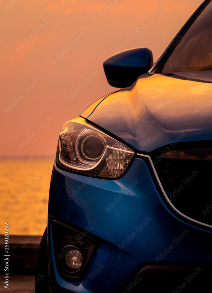 Front view of blue SUV car with sport and modern design parked on concrete road by the sea at sunset in the evening. Closeup headlamp light of blue car.  Hybrid and electric car technology concept.