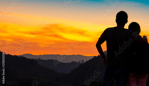 Back view of romantic couple watching beautiful sunset over mountain layer. Couple travel. Backpacker happy in love and travelling honeymoon. Silhouette of two tourist hiking to top of mountain.