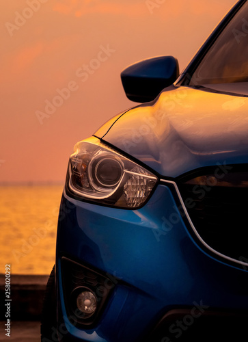 Front view of blue SUV car with sport and modern design parked on concrete road by the sea at sunset in the evening. Closeup headlamp light of blue car. Hybrid and electric car technology concept. © Artinun