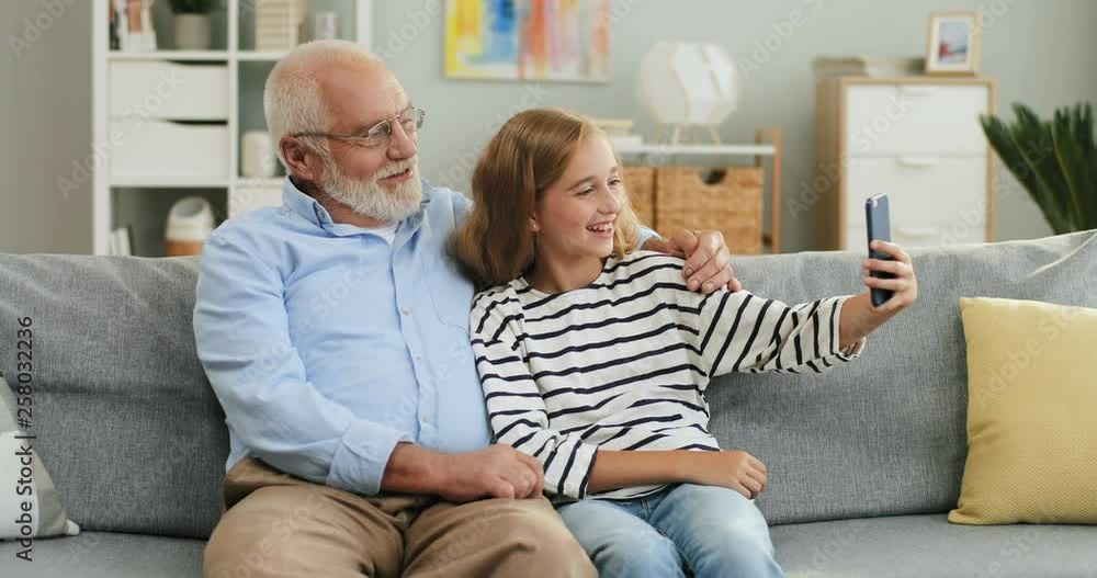Portrait shot of the happy smiled Caucasian grandfather and teenage granddaughter taking selfie photos while sitting on the couch at home. Portrait shot of the happy smiled Caucasian grandfather and teenage granddaughter taking selfie photos while sitting on the couch at home.