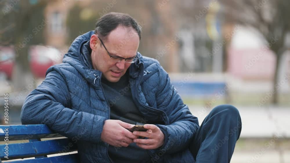 bored man in glasses siting on a bench and  using smartphone on the street in slow motion