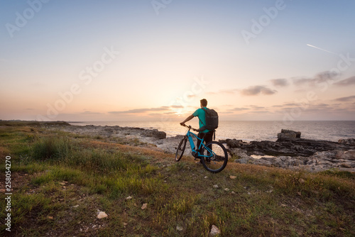 Man with a bike in the nature / Rear view of a man with a backpack and a bike enjoys the view of sunrise at the Black Sea coast