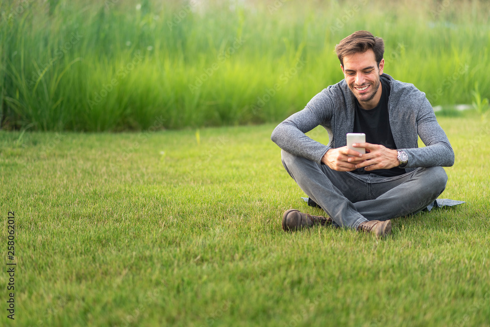 Young man sitting in the park, enjoying a nice conversation over his cell phone, surrounded by green grass; casual scene Young man sitting in the park, enjoying a nice conversation over his cell phone, surrounded by green grass; casual scene