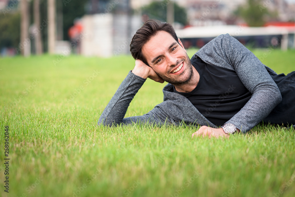 Young handsome man lying down on the green grass, enjoying a sunset relaxation in a park