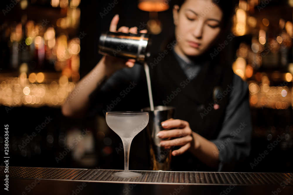 Bartender girl pouring a cocktail in steel shaker on the foreground of ice glass