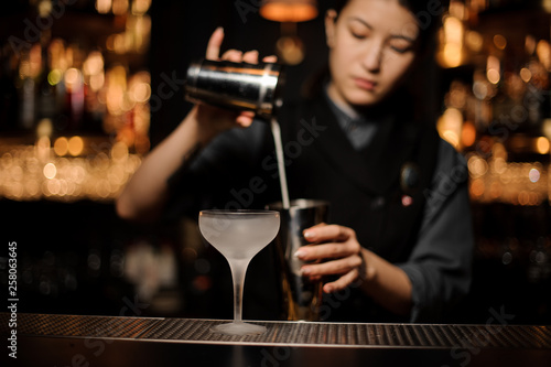Bartender girl pouring a cocktail in steel shaker on the foreground of ice glass