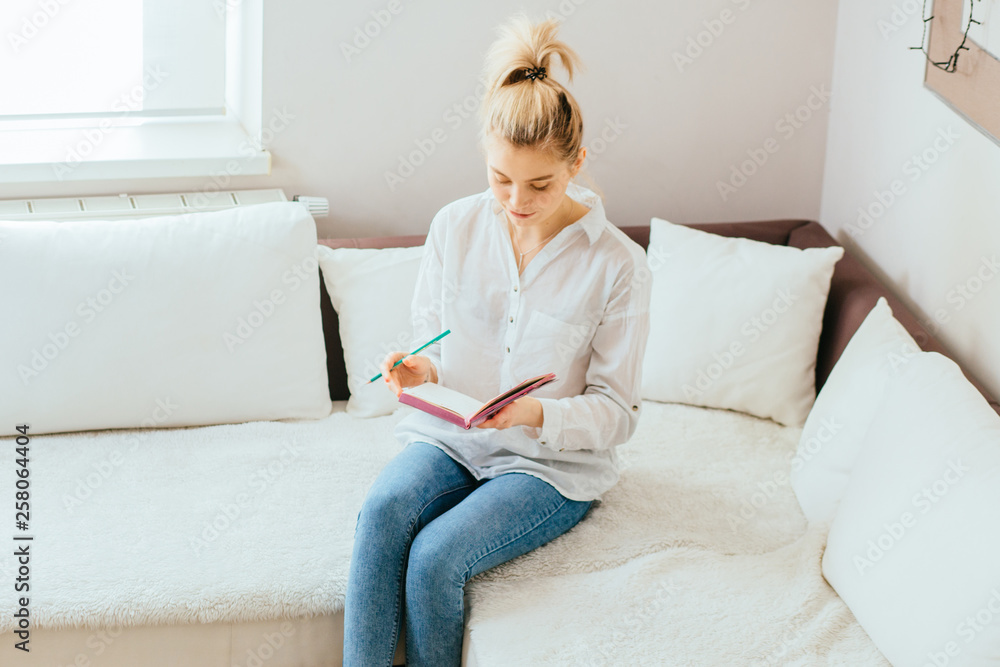 Portrait of young happy blond woman in blue jeans white shirt writing on paper, sitting on sofa at living room interior. Portrait of young happy blond woman in blue jeans white shirt writing on paper, sitting on sofa at living room interior.