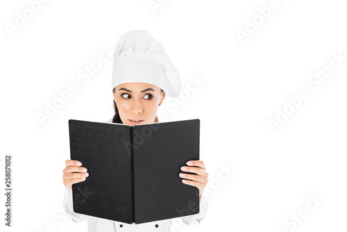 Curious chef in uniform holding black book isolated on white