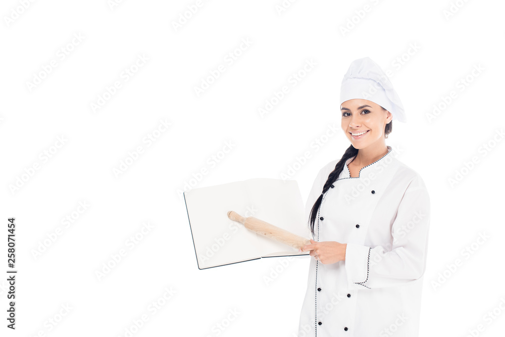 Smiling chef in hat holding open book and rolling pin isolated on white Smiling chef in hat holding open book and rolling pin isolated on white