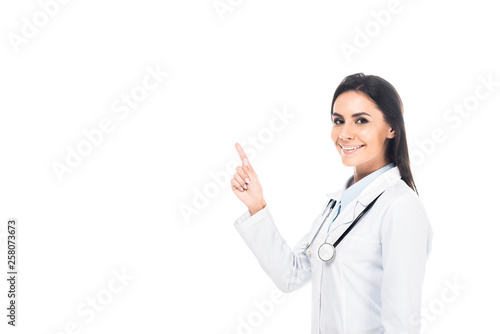 Smiling doctor in white coat with stethoscope pointing with finger isolated on white