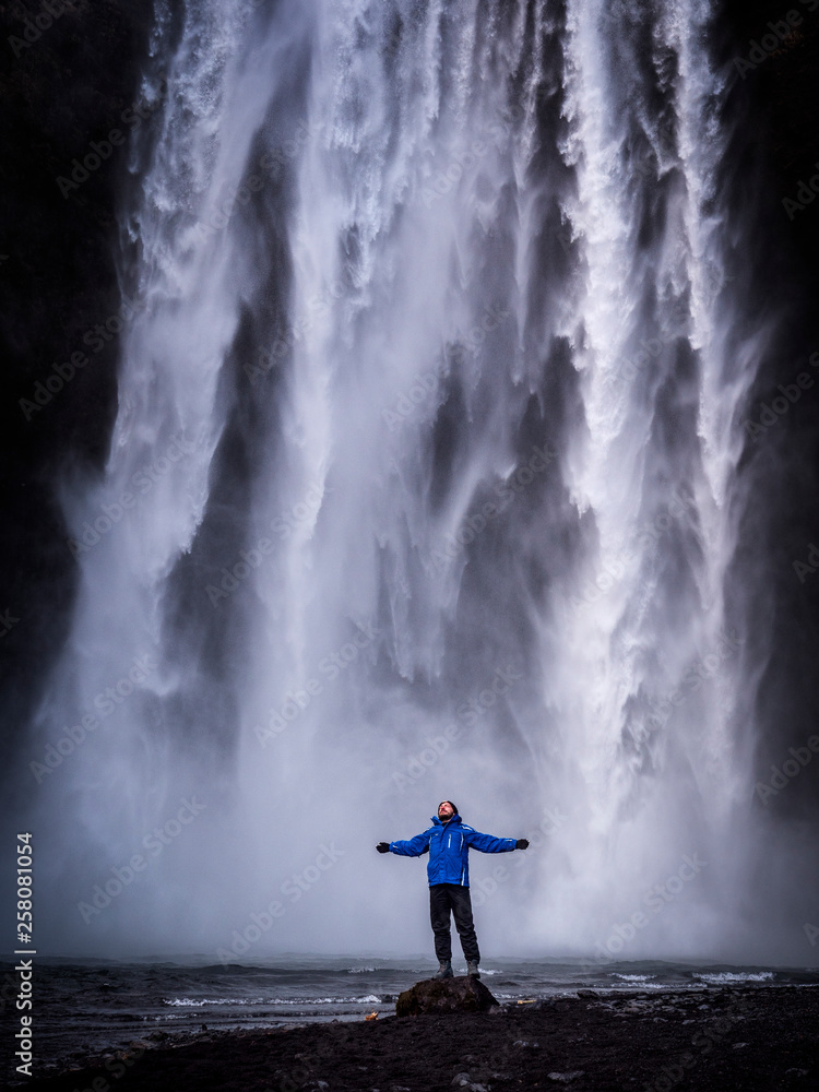 Obraz premium Male tourist in warm clothes stretching out arms and standing near Skogafoss Waterfall in Iceland