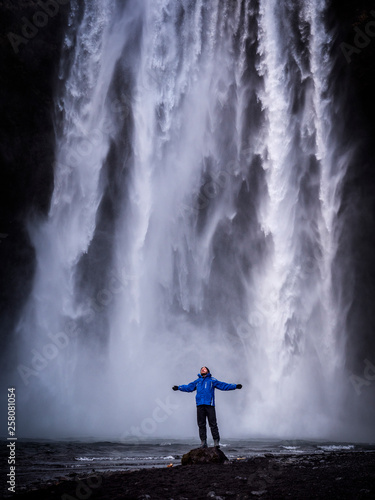 Male tourist in warm clothes stretching out arms and standing near Skogafoss Waterfall in Iceland