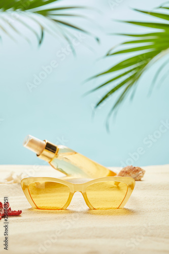 selective focus of sunglasses near bottle with suntan oil on sand isolated on blue