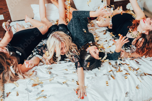 Theme party. Chill out. Group of girls in black relaxing on bed under confetti rain. BFF female gathering excitement.