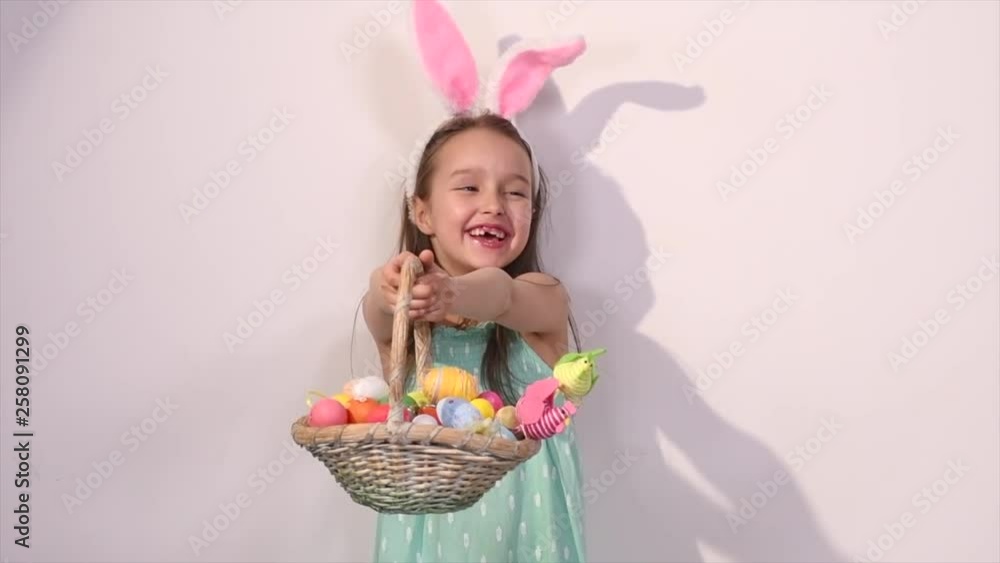 Cute little child wearing bunny ears on Easter day. Smiling happy Girl holding basket with painted colorful eggs and smiling. Happy spring holiday. Traditional colorful eggs. Slow motion 4K UHD video Cute little child wearing bunny ears on Easter day. Smiling happy Girl holding basket with painted colorful eggs and smiling. Happy spring holiday. Traditional colorful eggs. Slow motion 4K UHD video