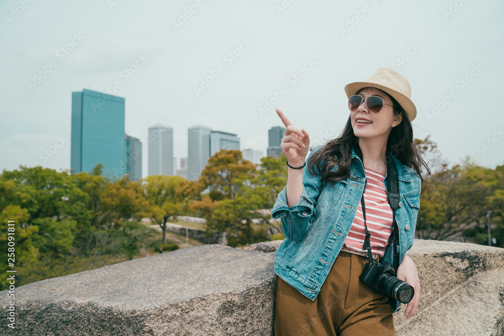 Young korean woman tourist leaning standing on top of rock in outdoor spring park osaka japan. girl pointing finger to sky with background skyscraper over big city urban view with tree in wild nature