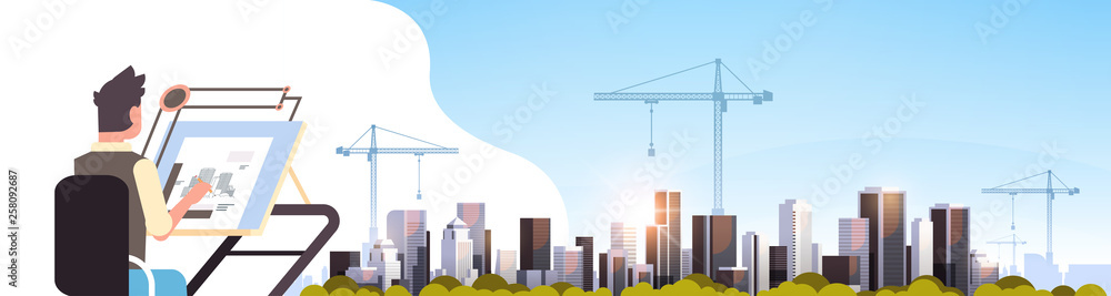 architect drawing blueprint urban building plan on adjustable board over city construction site tower cranes residential buildings cityscape sunset skyline background horizontal portrait