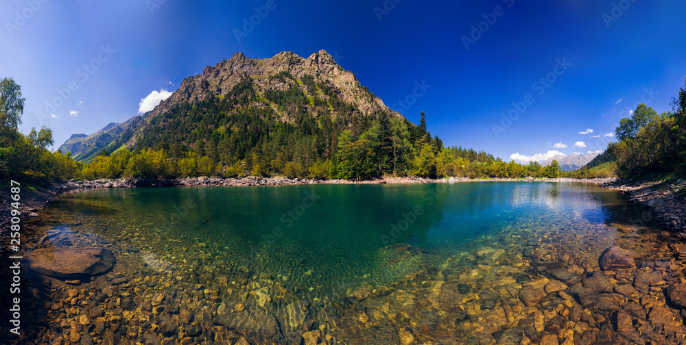 A huge panorama of the High-mountainous Baduk lake among the high Caucasian mountains and beautiful trees. Sunny summer day.