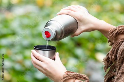 Woman pouring a hot drink in mug from thermos, and drinking tea during hike.