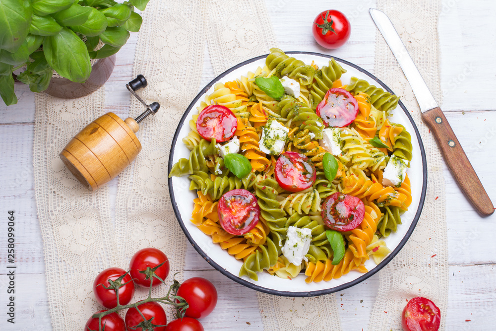 Italian food - Salad with colorful pasta, cherry tomatoes, feta cheese and fresh basil on white wooden background Italian food - Salad with colorful pasta, cherry tomatoes, feta cheese and fresh basil on white wooden background